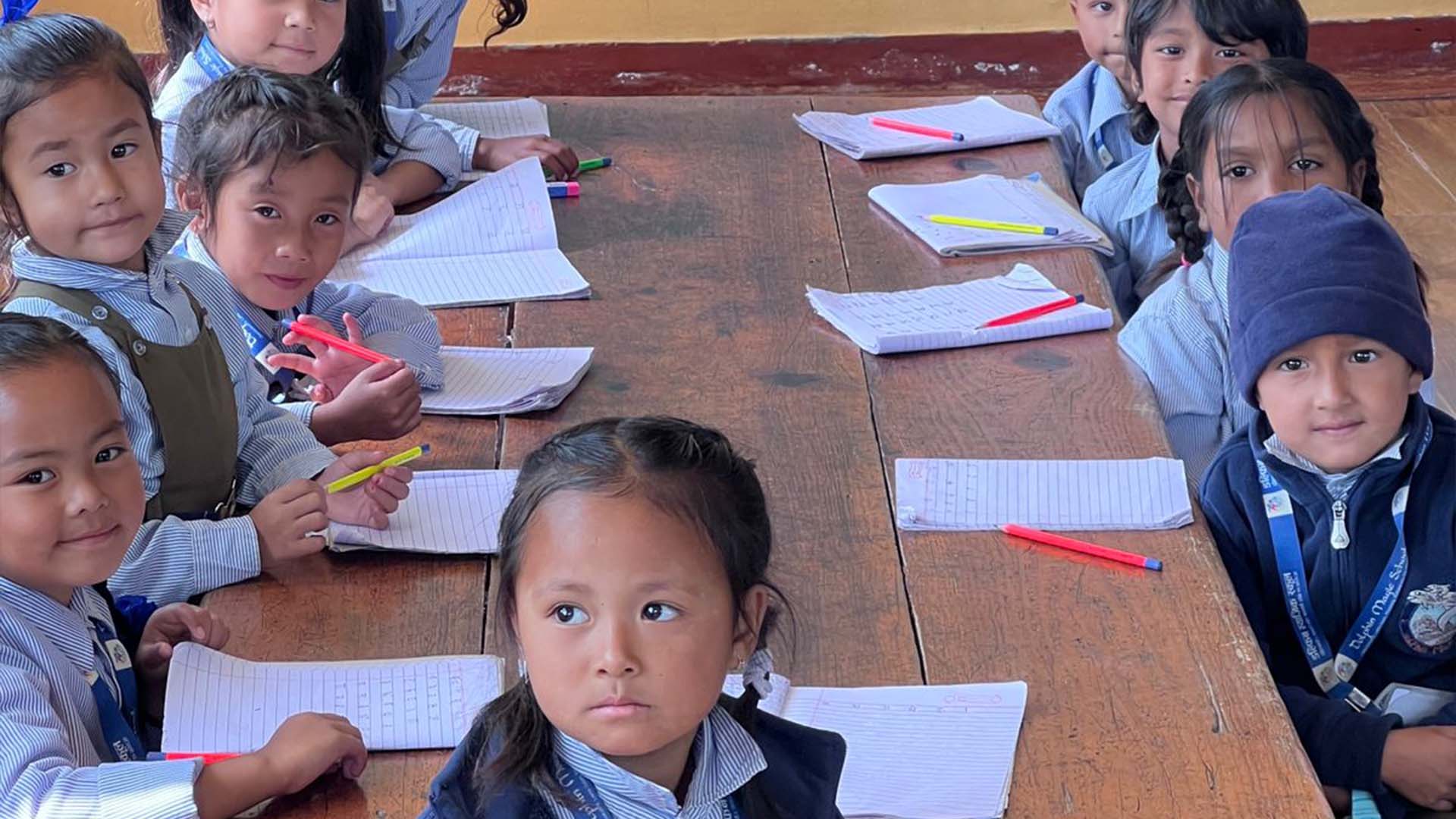 Mehrere Kinder sitzen an einem langen Tisch mit Schreibutensilien vor sich und schauen in die Kamera